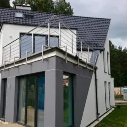 Dwukondygnacyjny dom z białymi ścianami i szarym dobudowanym segmentem, z metalową balustradą na dachu dobudówki, widoczne drzewa w tle i elementy placu budowy.