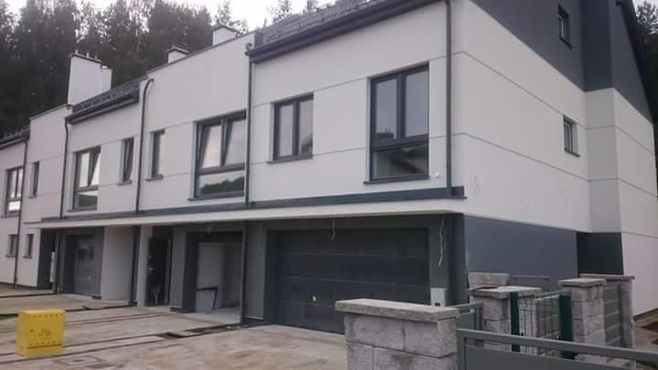 Nowoczesne szeregowe domy z jasną elewacją i ciemnymi akcentami, widok z zewnątrz, z częściowo widocznym ogrodzeniem i żółtym elementem na podjeździe.