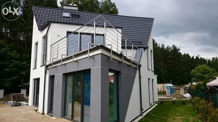 Dwukondygnacyjny dom z białymi ścianami i szarym dobudowanym segmentem, z metalową balustradą na dachu dobudówki, widoczne drzewa w tle i elementy placu budowy.