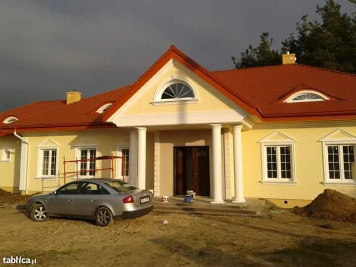 Żółty dom jednorodzinny z czerwonym dachem i białymi kolumnami przy wejściu, częściowo ocieplony, z samochodem osobowym zaparkowanym obok.