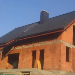 Dom w stanie surowym otwartym z ciemną dachówką i widocznym kominem, z elementami drewnianymi konstrukcji dachu, cegły porotherm.