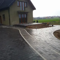 Nowoczesna posesja z ciemnoszarą kostką brukową ułożoną w łuki, z jasnymi pasami dekoracyjnymi, widoczni dwaj pracownicy na tarasie, dom w tle w trakcie wykończenia.