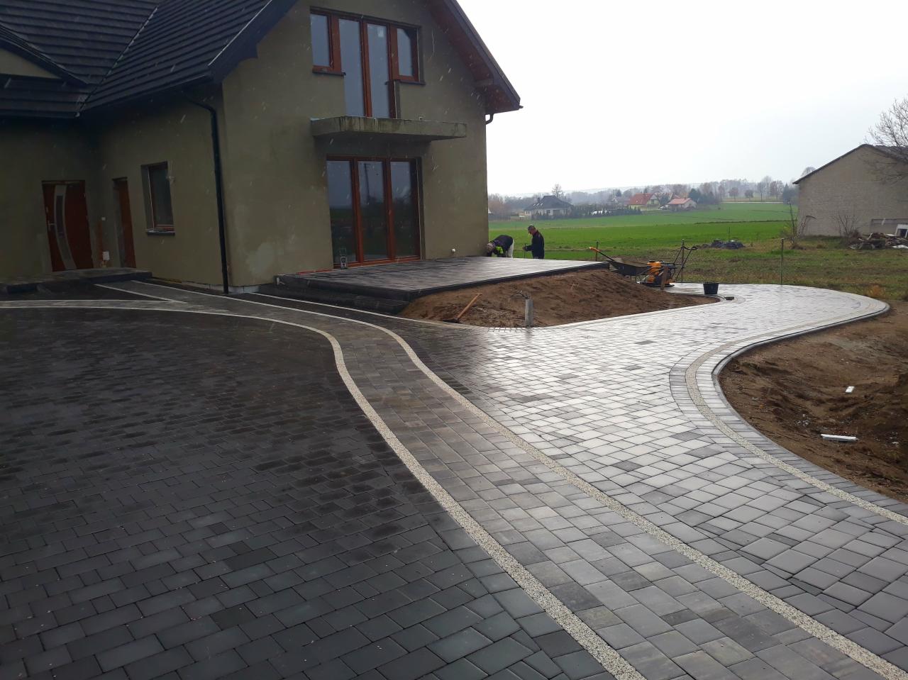 Nowoczesna posesja z ciemnoszarą kostką brukową ułożoną w łuki, z jasnymi pasami dekoracyjnymi, widoczni dwaj pracownicy na tarasie, dom w tle w trakcie wykończenia.