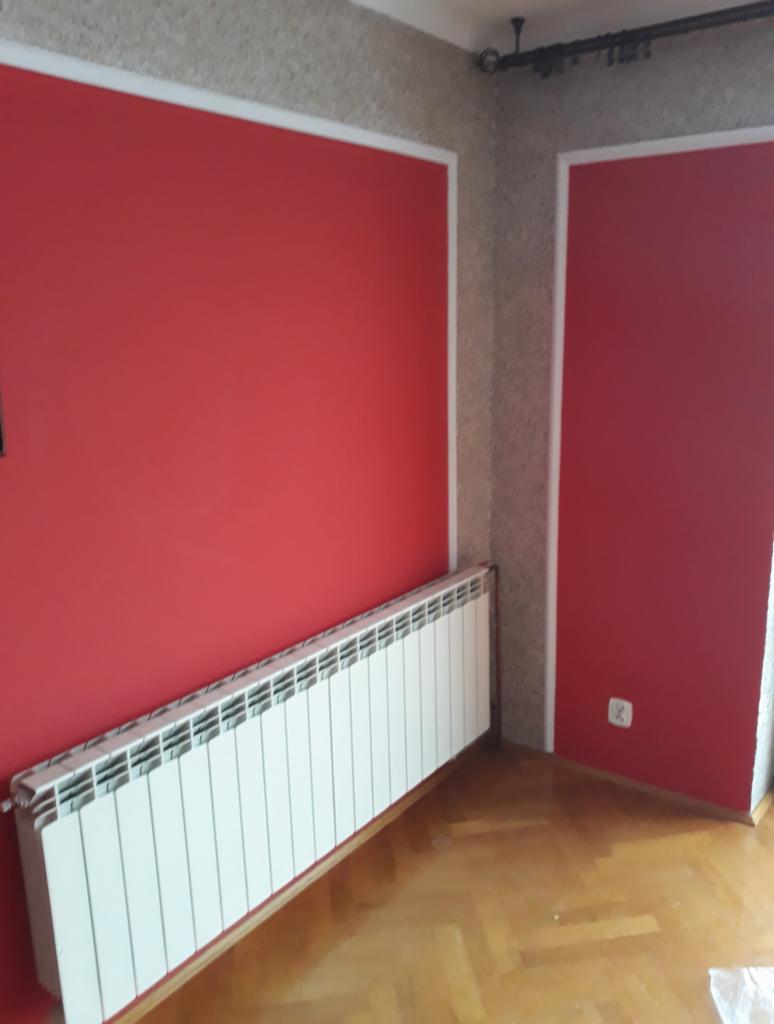 Czerwona ściana z białym obramowaniem, biały grzejnik żeberkowy i parkiet w jodełkę w pustym pokoju po remoncie.