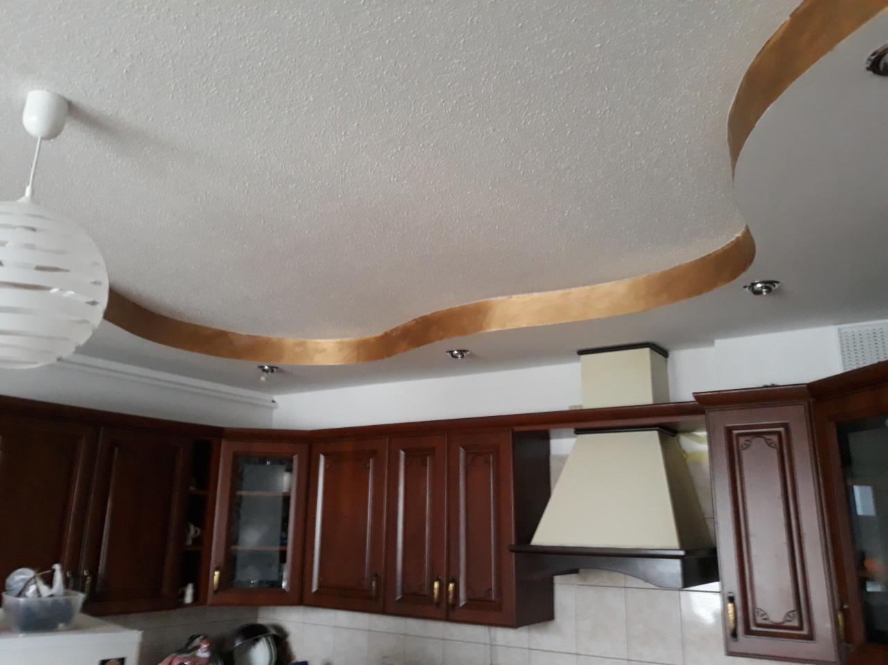 Sufit w kuchni z dekoracyjnym, falistym obramowaniem w kolorze złota, widoczne brązowe szafki kuchenne i lampa wisząca.
