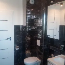 Nowoczesna łazienka z czarnymi marmurkowymi płytkami, lustrzaną mozaiką, podwieszaną toaletą i białą umywalką z szafką, widok na drzwi z poziomymi paskami.