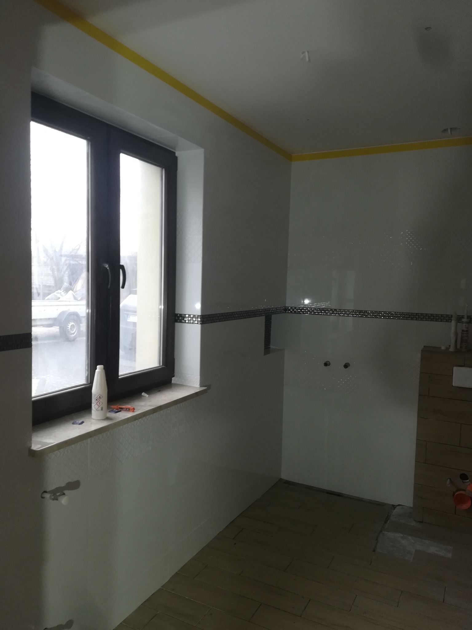 Wnętrze łazienki w trakcie prac wykończeniowych: położone płytki ścienne z dekoracyjnym pasem, drewnopodobne płytki podłogowe, okno z widokiem na zewnątrz, żółta taśma malarska na suficie.