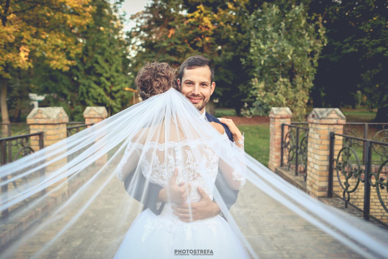Uśmiechnięty pan młody obejmuje pannę młodą w koronkowej sukni, welon rozciągnięty na moście z ceglanymi barierkami, drzewa w tle.