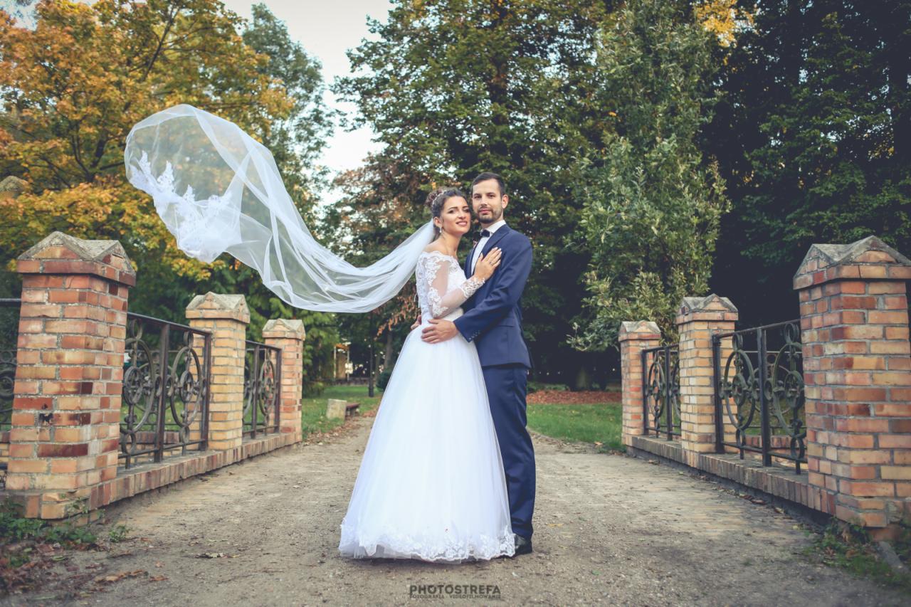 Para młoda na moście w Lesznie, panna młoda w białej sukni z długim welonem, pan młody w granatowym garniturze, jesienne drzewa w tle.