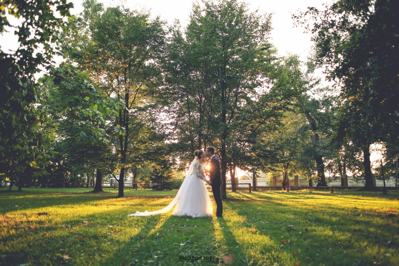 Para młoda całująca się na tle drzew w słoneczny dzień, długi welon panny młodej rozciąga się na trawie, promienie słoneczne przebijają przez liście.