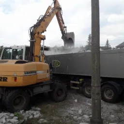 Żółta koparka Liebherr 314 ładuje gruz do szarej naczepy ciężarówki; w tle budynek mieszkalny i pochmurne niebo.