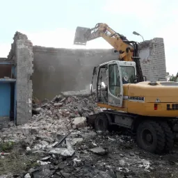 Żółta koparka Liebherr 314 z łyżką zawieszoną nad gruzami ceglanego budynku w trakcie rozbiórki, widoczne fragmenty ścian i niebieskie drzwi, pył w powietrzu.