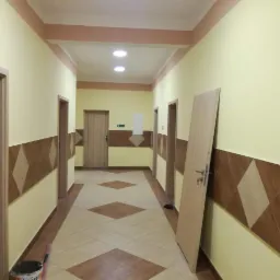 Korytarz po remoncie z żółtymi ścianami, brązowymi panelami ściennymi w romby i brązową podłogą. Przy ścianie oparta surowa, nielakierowana płyta drzwiowa z zamontowaną klamką i kluczem.