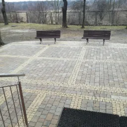 Plac wyłożony dwukolorową kostką brukową w kształcie kwadratów z ławkami, widok z góry z fragmentem metalowej barierki.