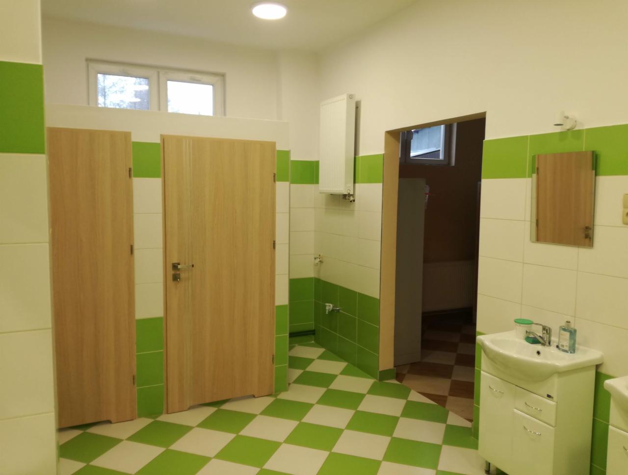 Wykończona łazienka publiczna z dwoma drewnianymi kabinami WC, umywalkami, lustrem i podłogą w zielono-białą szachownicę.