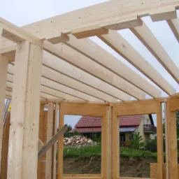 Konstrukcja drewniana domu szkieletowego w trakcie budowy, widoczne elementy nośne i więźba dachowa, w tle dom z czerwonym dachem i stos drewna.