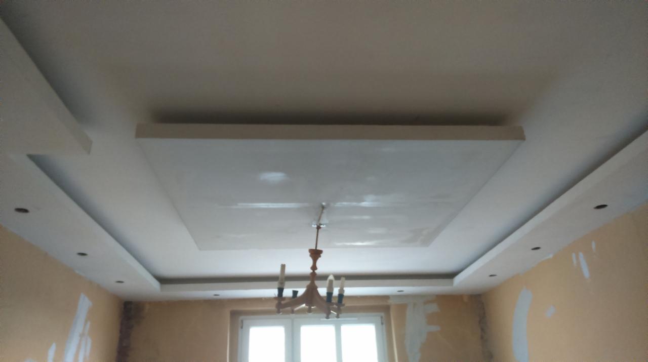 Wykończony sufit z obniżonym fragmentem w kształcie kwadratu, oświetlenie punktowe w listwach po bokach, ściany w trakcie malowania, widoczny żyrandol.