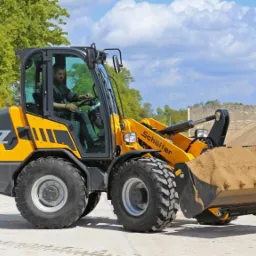 Żółta miniładowarka Schäffer 5470Z z operatorem, nabierająca piasek, na tle drzew i błękitnego nieba z chmurami.