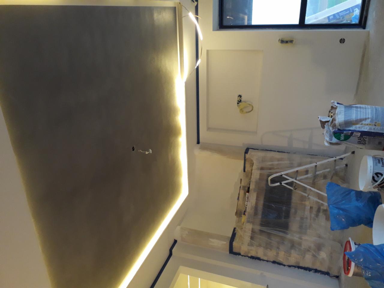 Pokój w trakcie remontu: świeżo pomalowany sufit z oświetleniem LED, zabezpieczone folią drzwi, drabina, worki z gipsem i puszki z farbą.