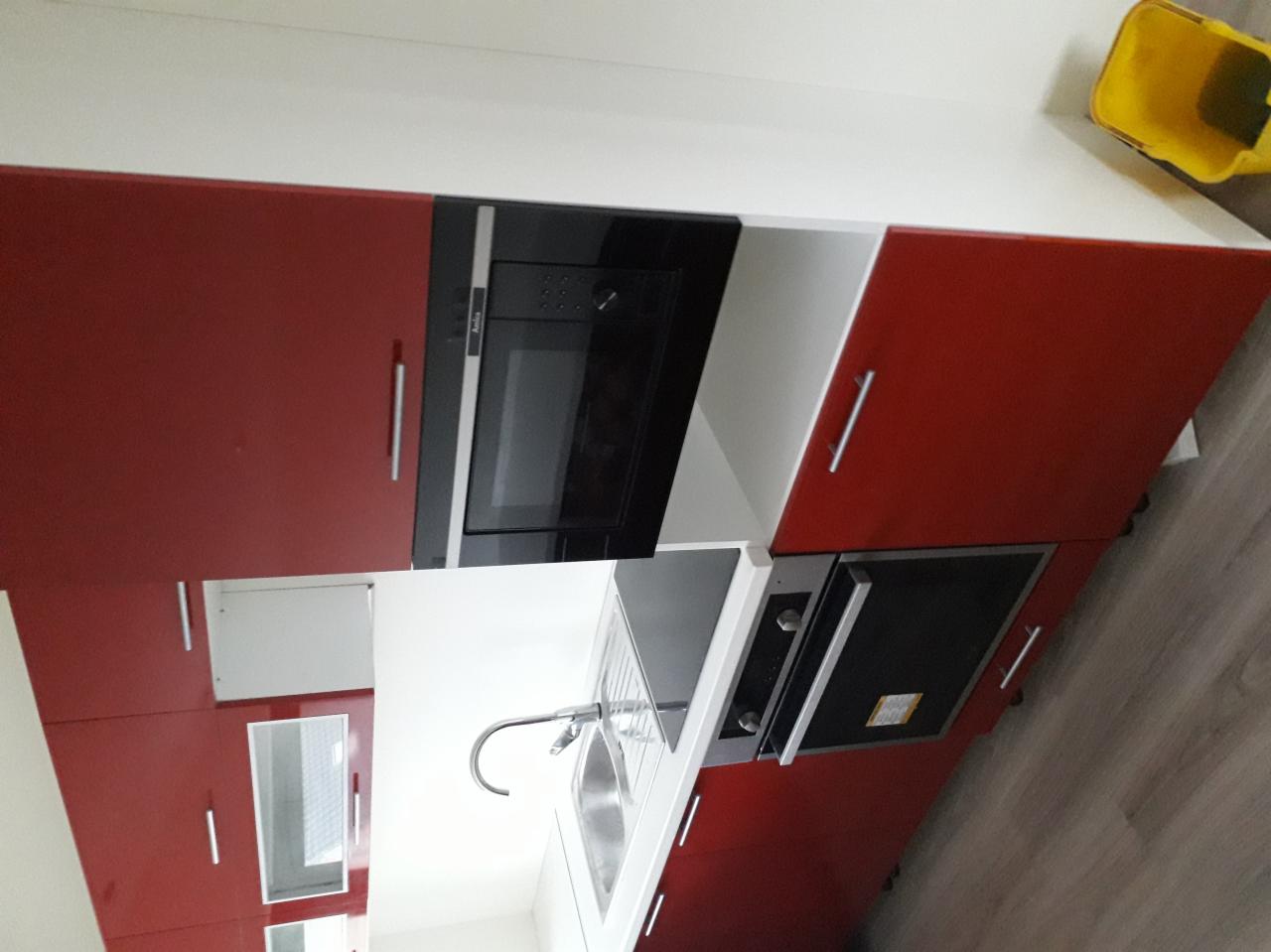 Nowoczesna kuchnia z czerwonymi szafkami, wbudowanym piekarnikiem i mikrofalówką, oraz stalowym zlewozmywakiem z baterią.