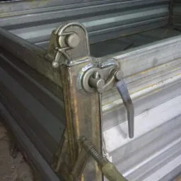 Fragment metalowej konstrukcji przyczepy z widocznym mechanizmem blokującym burtę, spawany element z dźwignią. Widoczne ślady spawania i obróbki metalu.