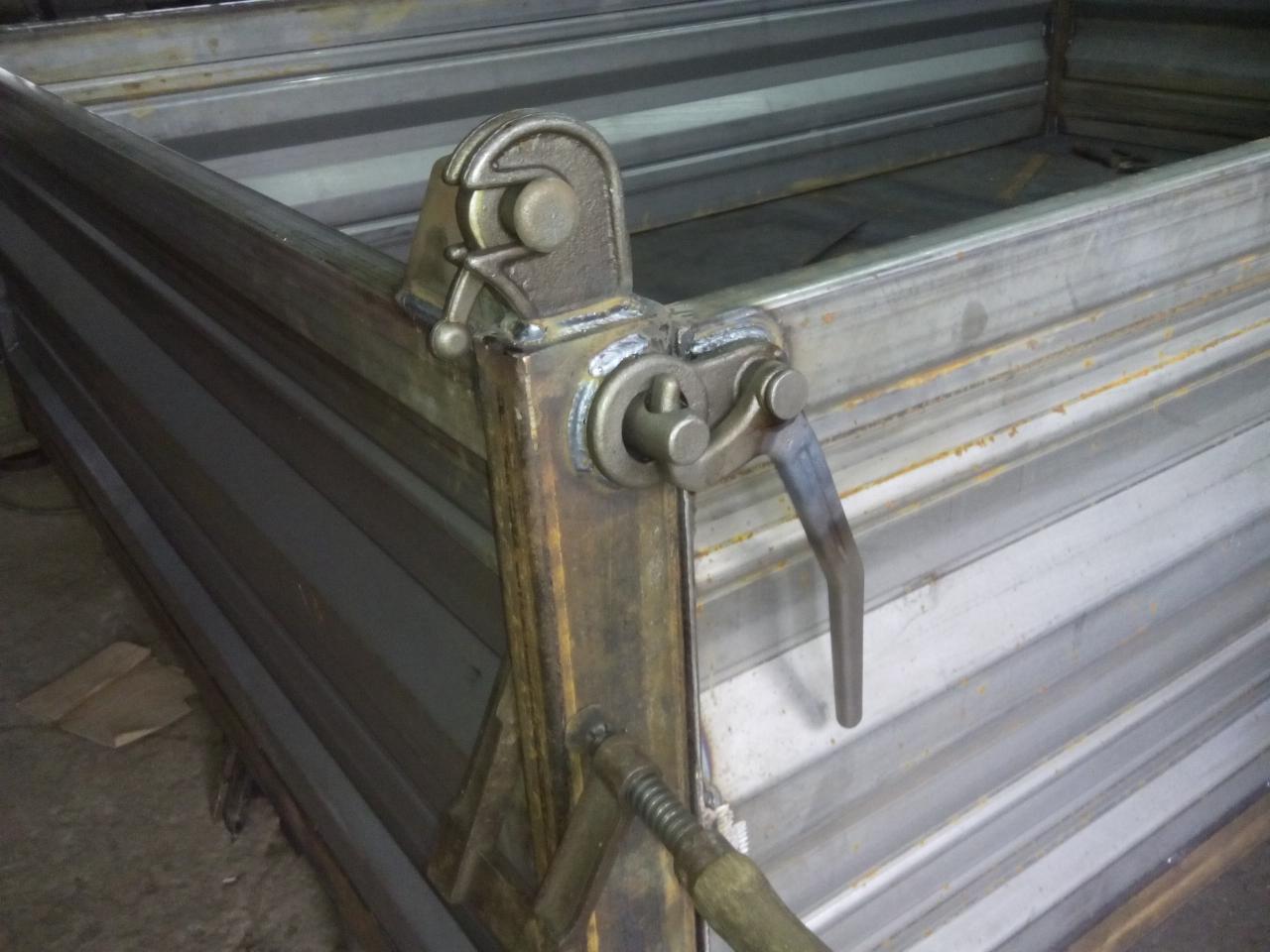 Fragment metalowej konstrukcji przyczepy z widocznym mechanizmem blokującym burtę, spawany element z dźwignią. Widoczne ślady spawania i obróbki metalu.