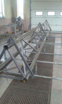 Konstrukcja kratownicowa z rur i profili stalowych, spawana, leżąca na metalowej siatce w warsztacie, widoczne ślady spawania.
