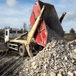 Ciężarówka z wywrotką w trakcie wysypywania gruzu budowlanego na placu, widoczny mechanizm wywrotki i łańcuchy zabezpieczające.