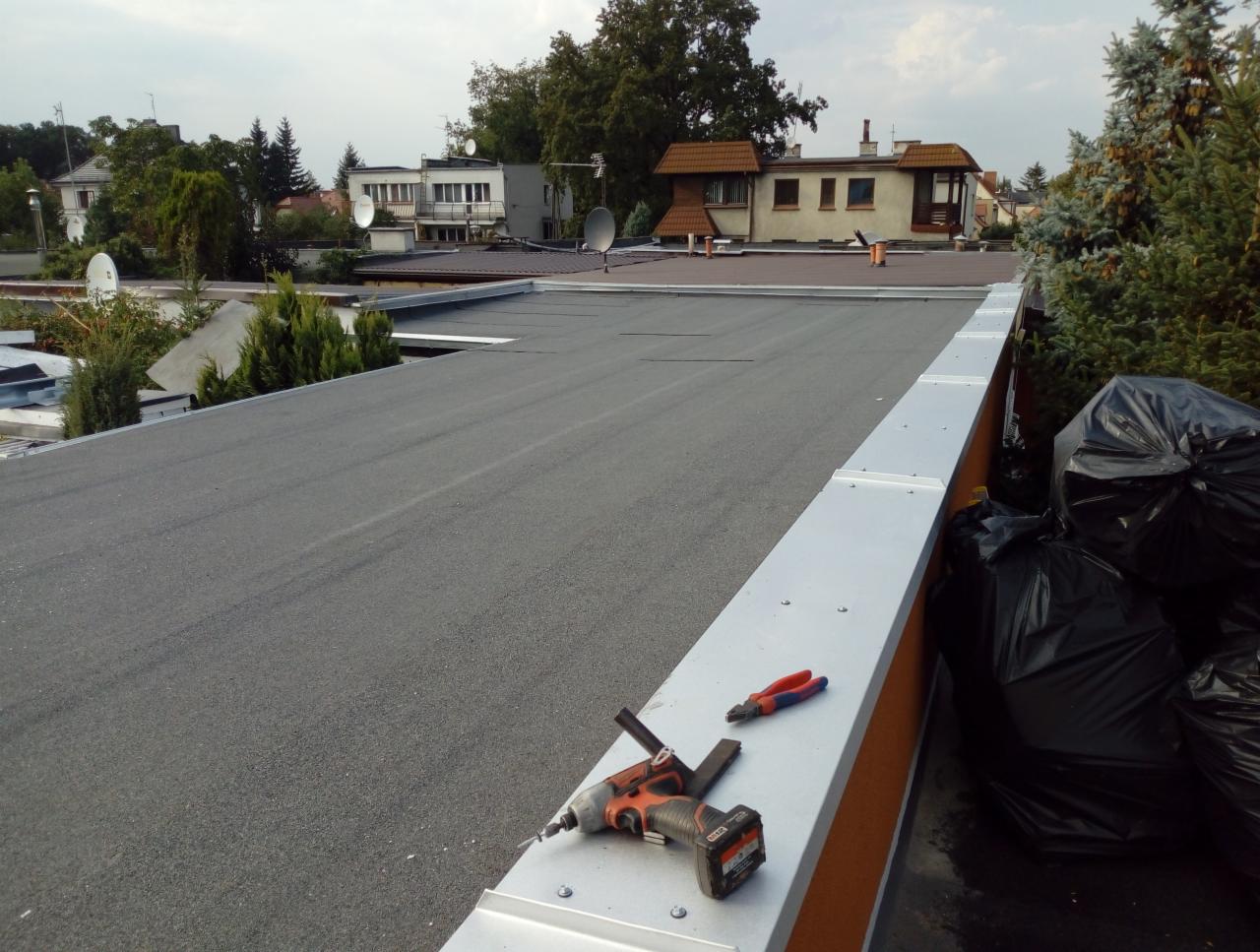 Czarny, płaski dach pokryty papą termozgrzewalną z widocznymi narzędziami: wkrętarką akumulatorową i kombinerkami, na tle zabudowań miejskich i drzew.