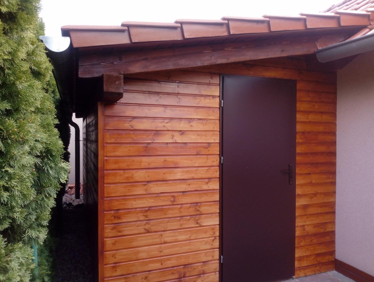 Drewniana wiata z brązowymi drzwiami i rynną, widok z boku, obok rosnący krzew