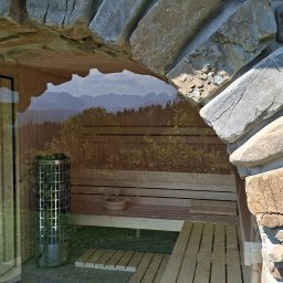 EMEN Maciej Niemczyk - Rustykalna sauna z kamiennym łukiem, widok na góry przez szybę. Wnętrze z drewnianymi ławkami i piecem. Elementy wystroju: drewniane wiadro i czerpak.