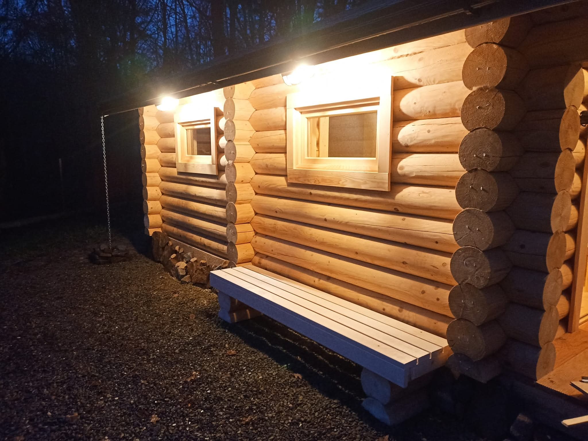 Zewnętrzny widok sauny z bali drewnianych oświetlonej lampami, z ławką przed wejściem, na tle ciemnego otoczenia. Widoczne detale konstrukcji i naturalne wykończenie.