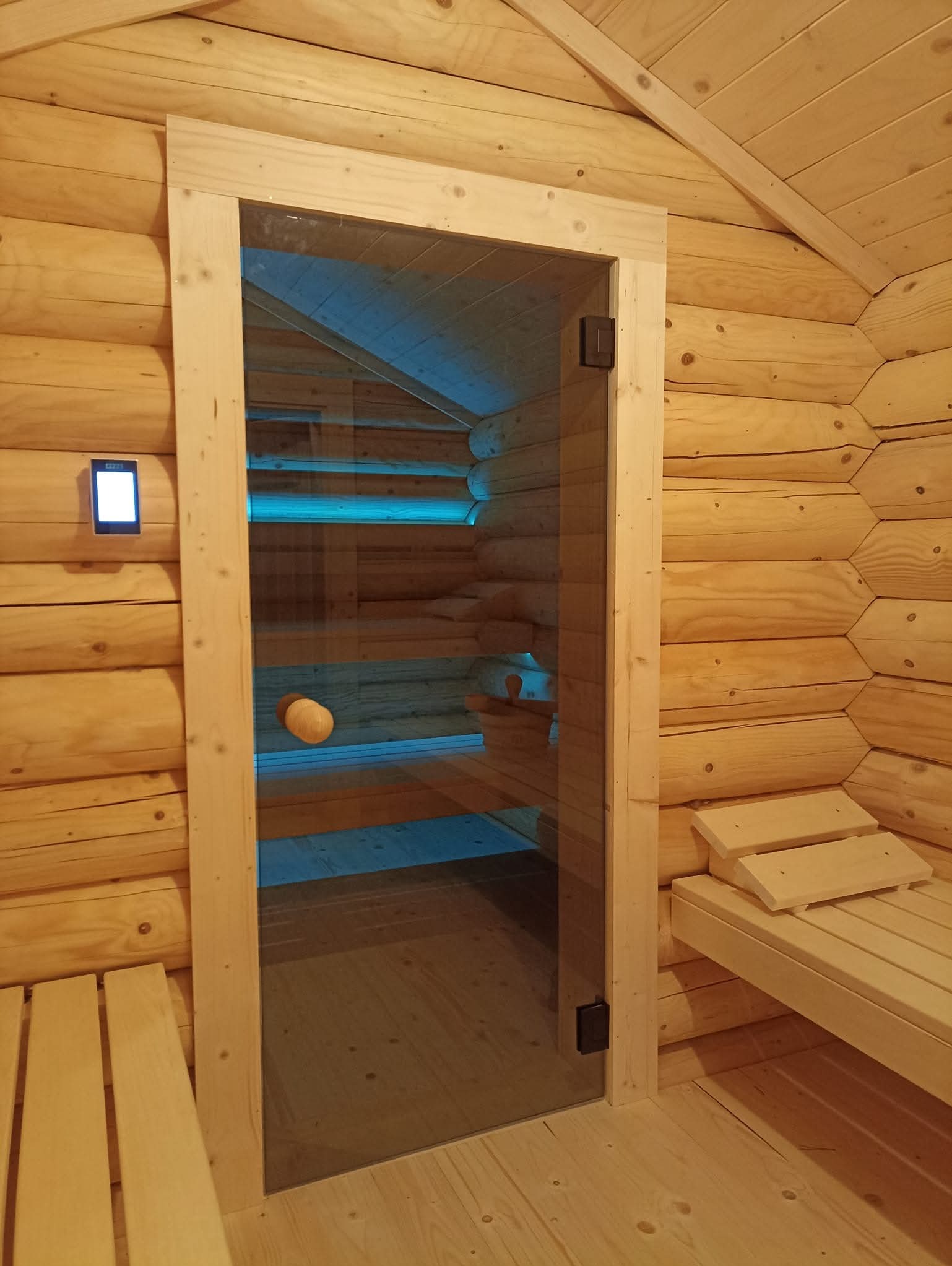 Wnętrze sauny z drewnianymi ławkami i szklanymi drzwiami, podświetlone niebieskim światłem LED. Na ścianie panel sterowania. Wykończenie z jasnego drewna.