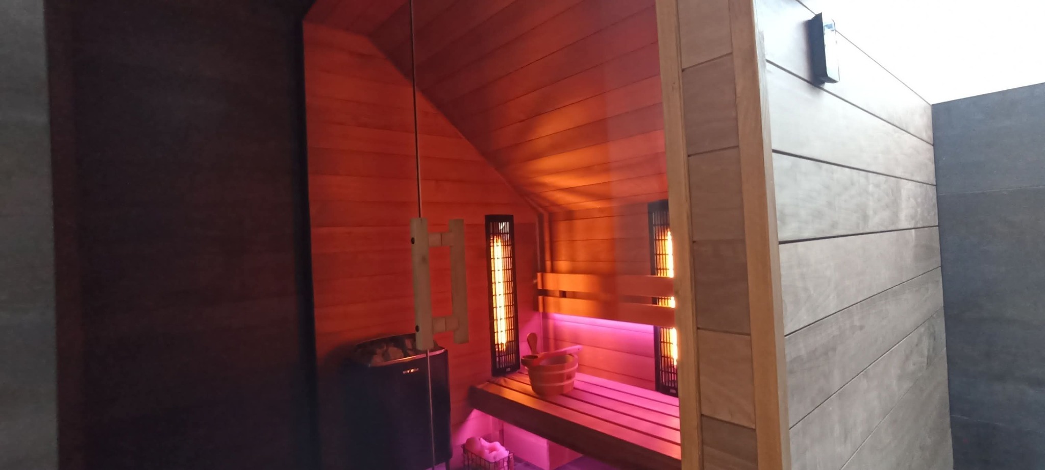 Nowoczesna sauna z drewnianym wykończeniem, oświetleniem LED i piecem. Widoczne detale: kubełek, chochla i panele grzewcze. Ujęcie z perspektywy wejścia.