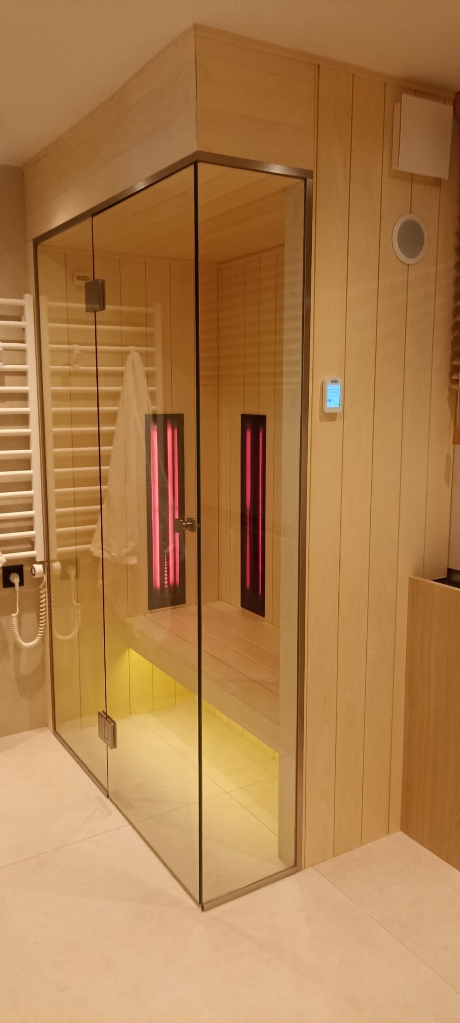 Nowoczesna sauna z przeszklonymi drzwiami i podświetleniem LED, w jasnym pomieszczeniu z grzejnikiem drabinkowym i drewnianymi panelami ściennymi.