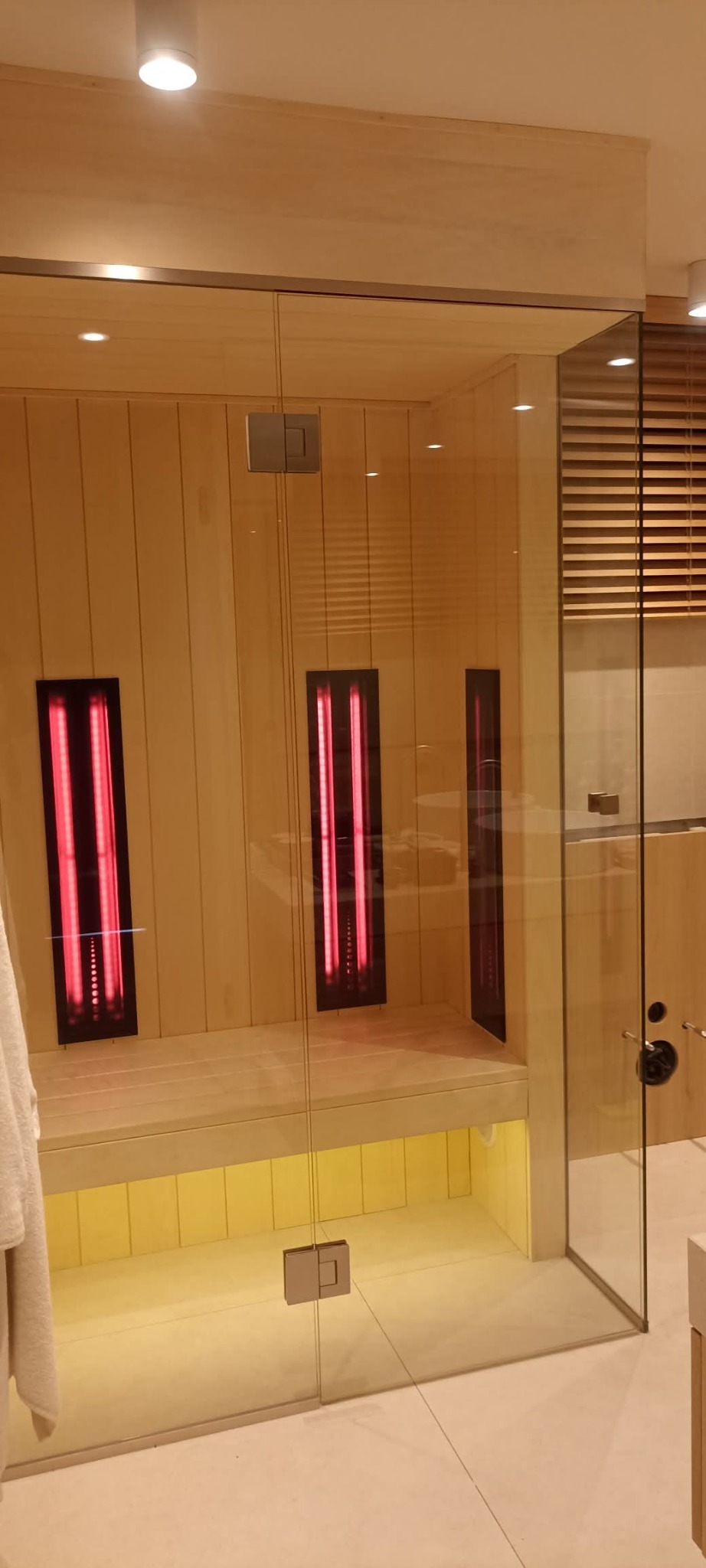 Nowoczesna sauna infrared z przeszklonymi drzwiami i drewnianym wnętrzem, oświetlona od dołu, z trzema panelami grzewczymi o czerwonym świetle.