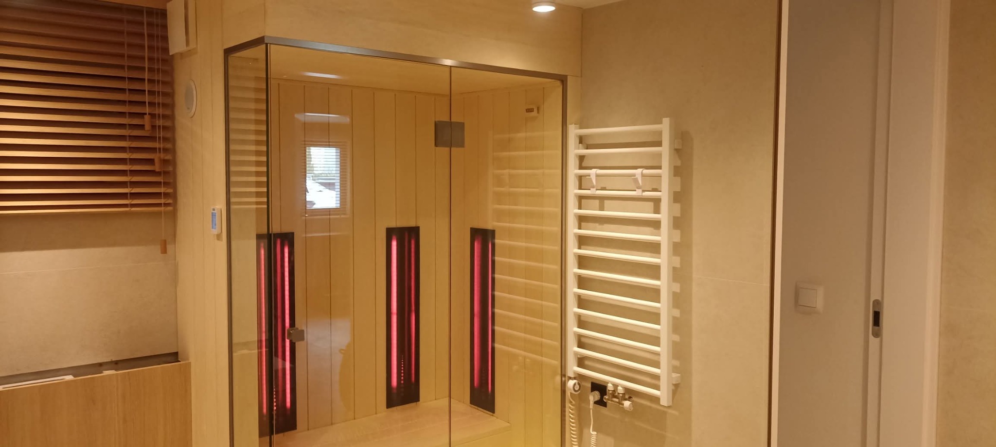 Nowoczesna sauna infrared z przeszklonymi ścianami i drewnianym wnętrzem, oświetlona czerwonymi panelami grzewczymi, w jasnej łazience z grzejnikiem drabinkowym.