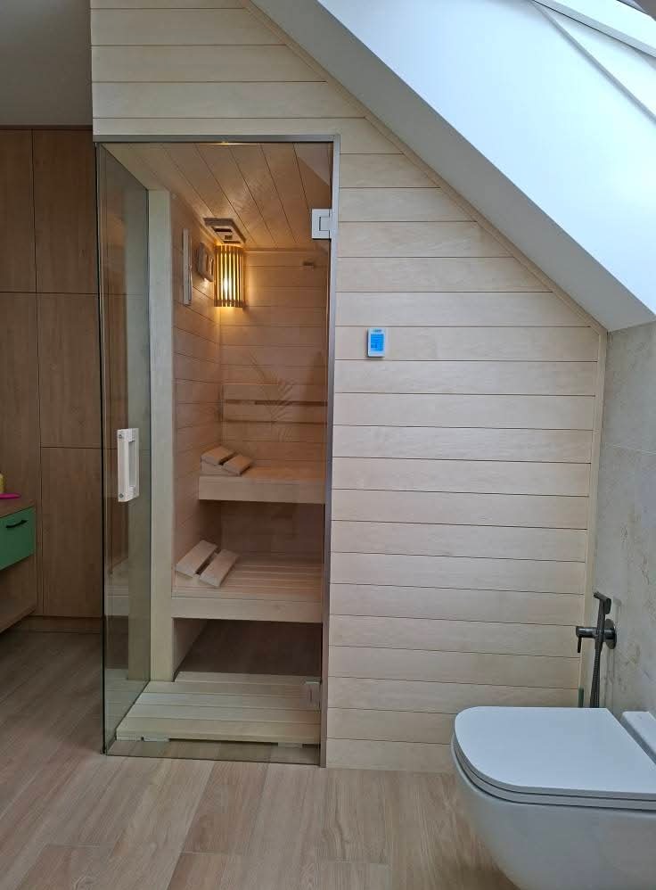Jasna, drewniana sauna wkomponowana w skos dachu, widoczna przez szklane drzwi. Wnętrze oświetlone lampą, z dwoma poziomami ławek. Obok toaleta i armatura.
