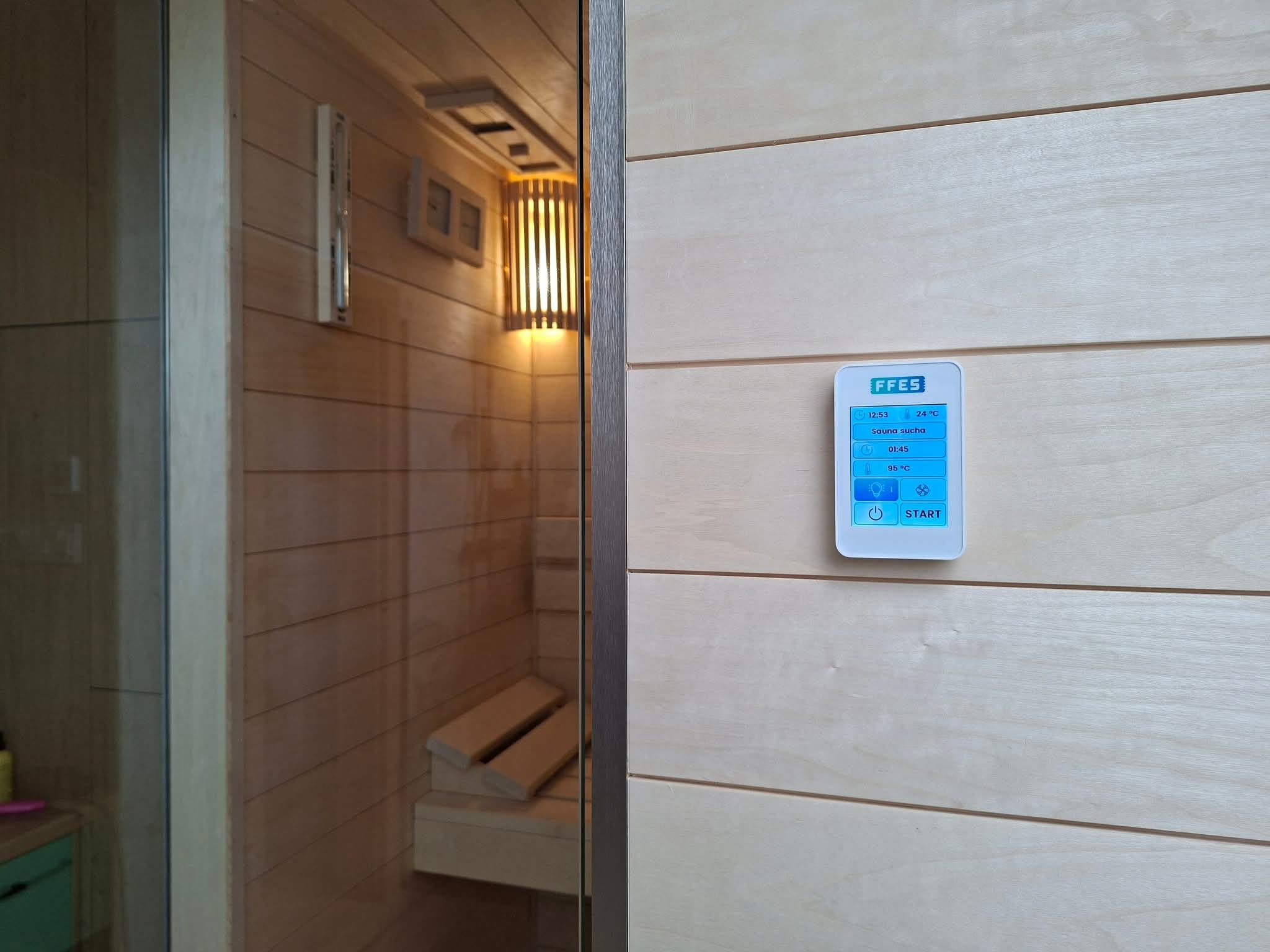 Panel sterowania sauny suchej z widoczną temperaturą 95°C, zamontowany na jasnej, drewnianej ścianie. W tle widoczne wnętrze sauny z oświetleniem i ławkami.