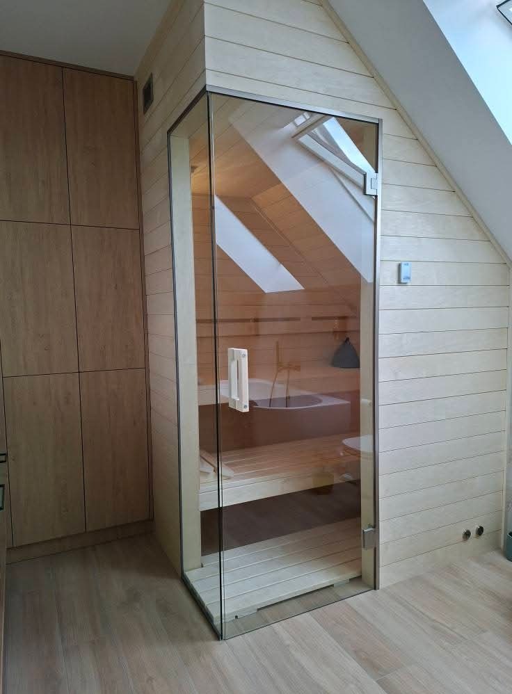 Jasna sauna w skandynawskim stylu z przeszklonymi drzwiami, widoczne drewniane ławki i okno dachowe. Minimalistyczne wykończenie wnętrza.