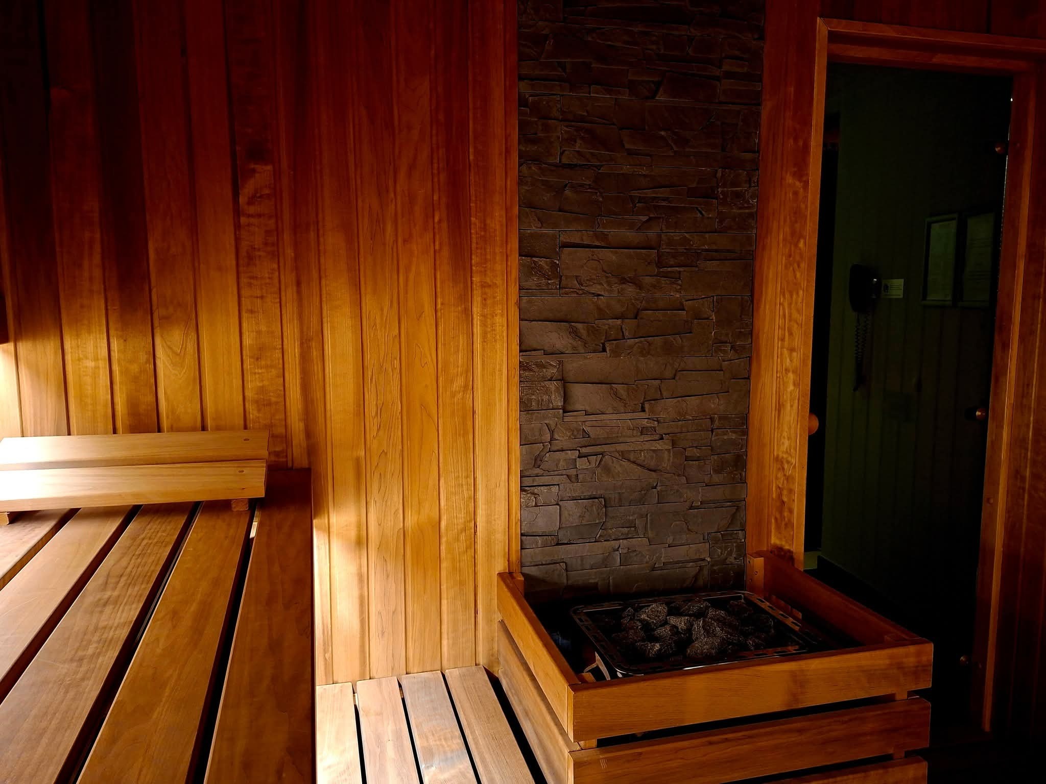 Wnętrze sauny z drewnianymi ławkami i piecem obłożonym kamieniami. Ściana wyłożona drewnem i kamieniem dekoracyjnym. Otwarta drewniana rama drzwi.