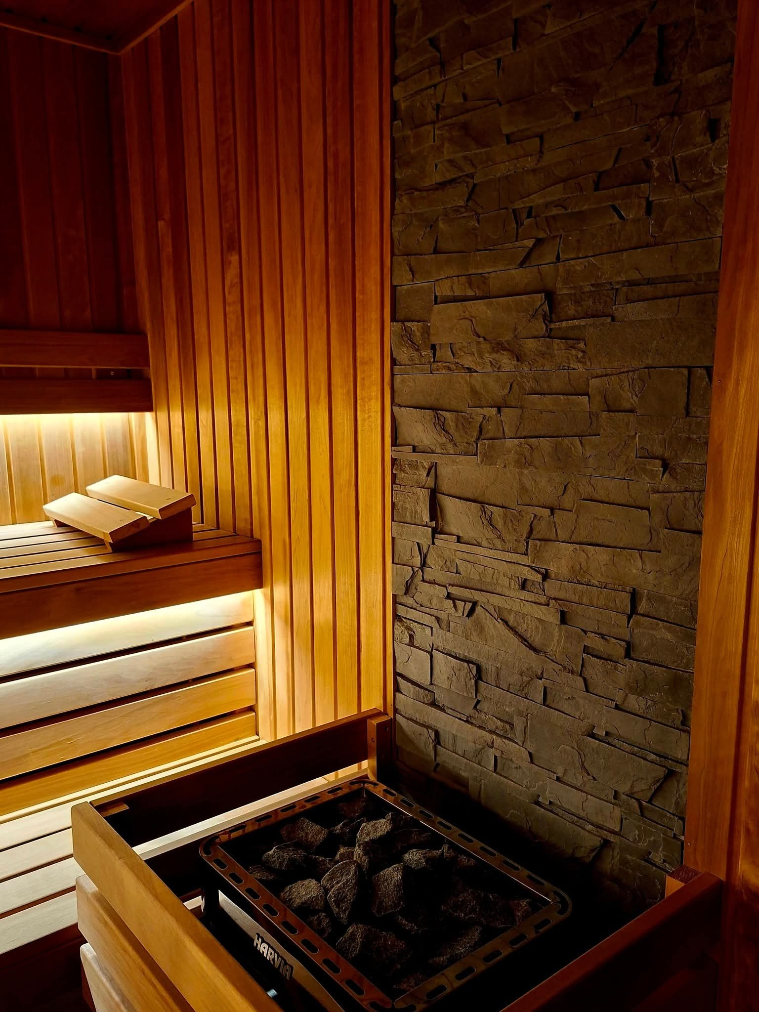 Drewniana sauna z piecem Harvia i kamienną ścianą. Podświetlone ławki z zagłówkami. Ciepłe, stonowane oświetlenie tworzy relaksującą atmosferę.