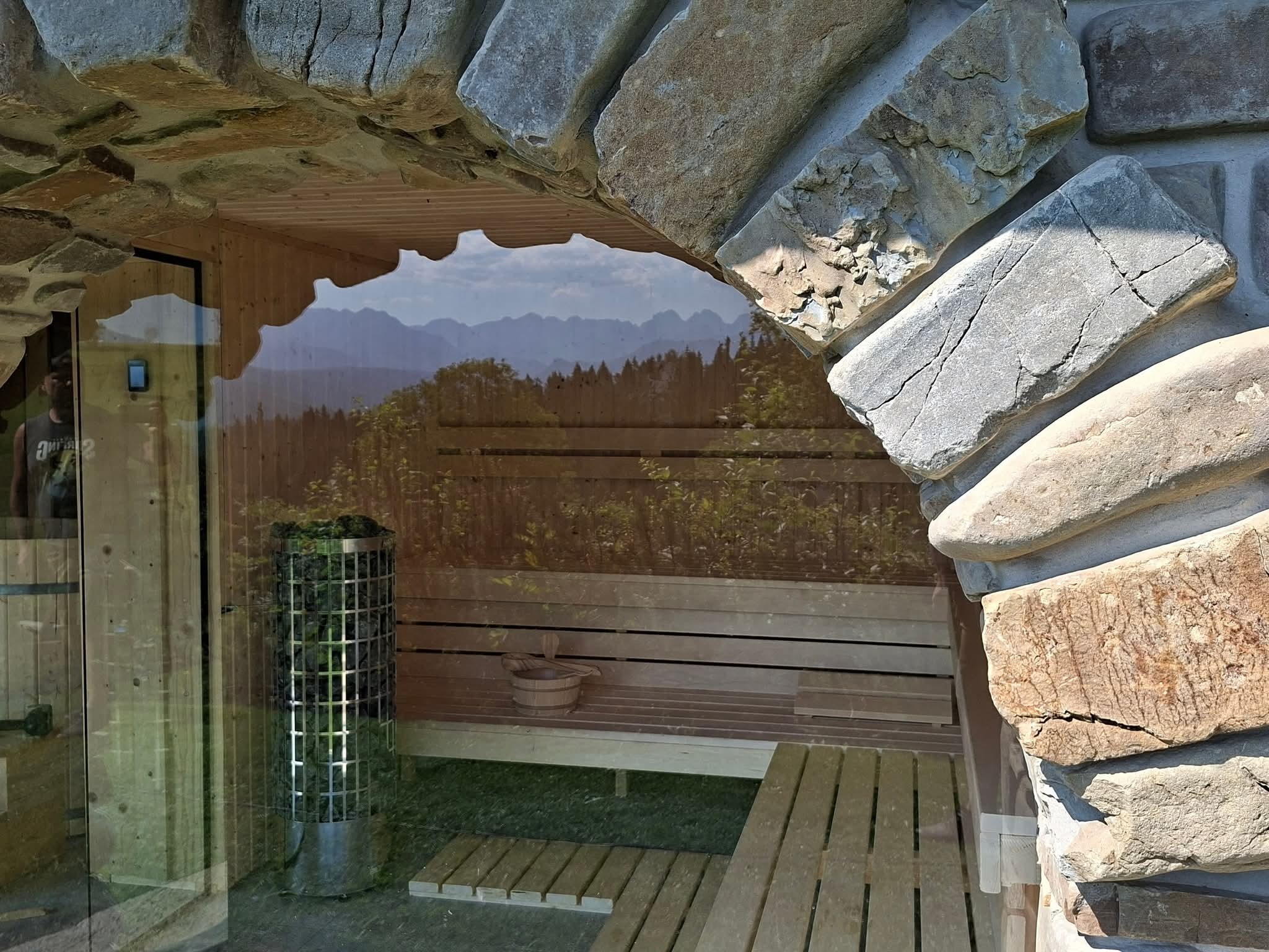 Rustykalna sauna z kamiennym łukiem, widok na góry przez szybę. Wnętrze z drewnianymi ławkami i piecem. Elementy wystroju: drewniane wiadro i czerpak.