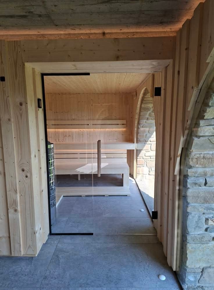 Nowoczesna sauna z przeszklonymi drzwiami i drewnianym wnętrzem, oświetlona subtelnym światłem. Widoczny piec i kamienna ściana obok wejścia.