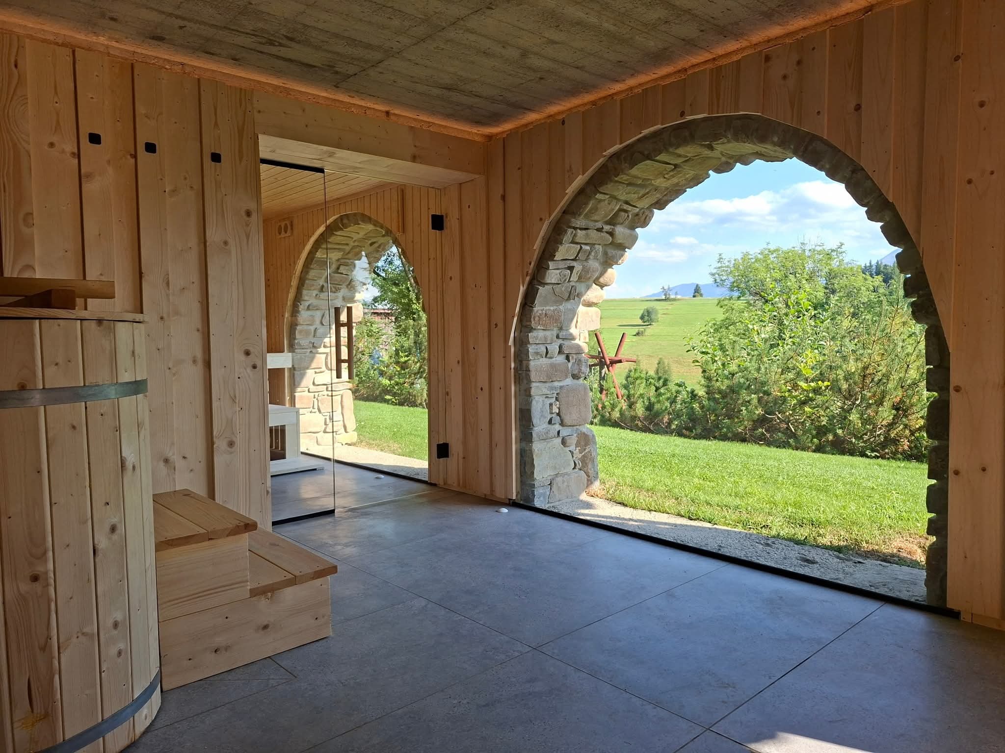 Drewniana sauna z kamiennymi łukami okiennymi, widok na zielone wzgórza. Wnętrze wykończone drewnem, z drewnianą beczką i schodkami. Nowoczesny design z naturalnymi elementami.
