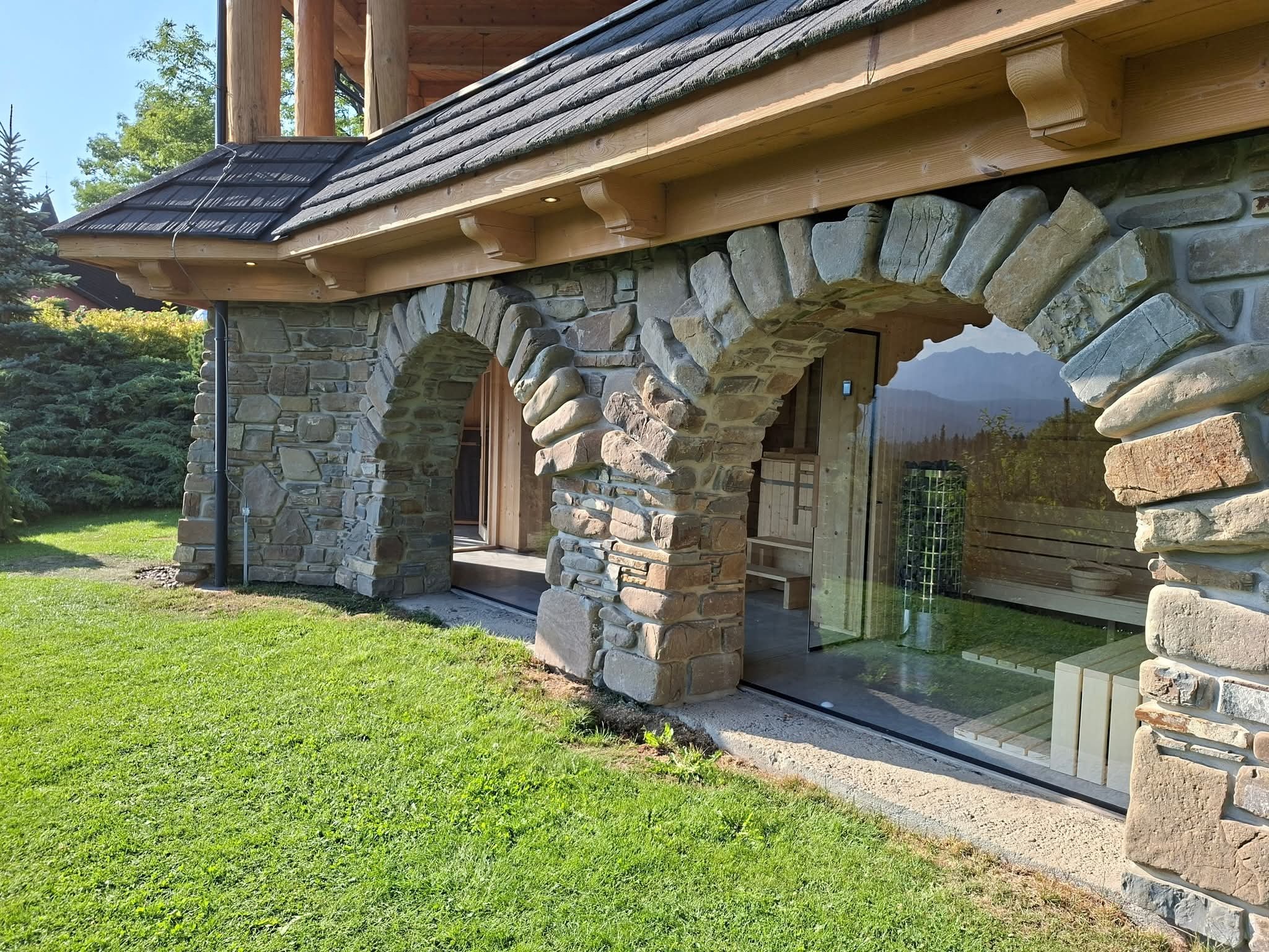 Kamienna sauna zewnętrzna z łukowym wejściem i widokiem na góry, otoczona zielonym trawnikiem. Detale architektoniczne z naturalnego kamienia.