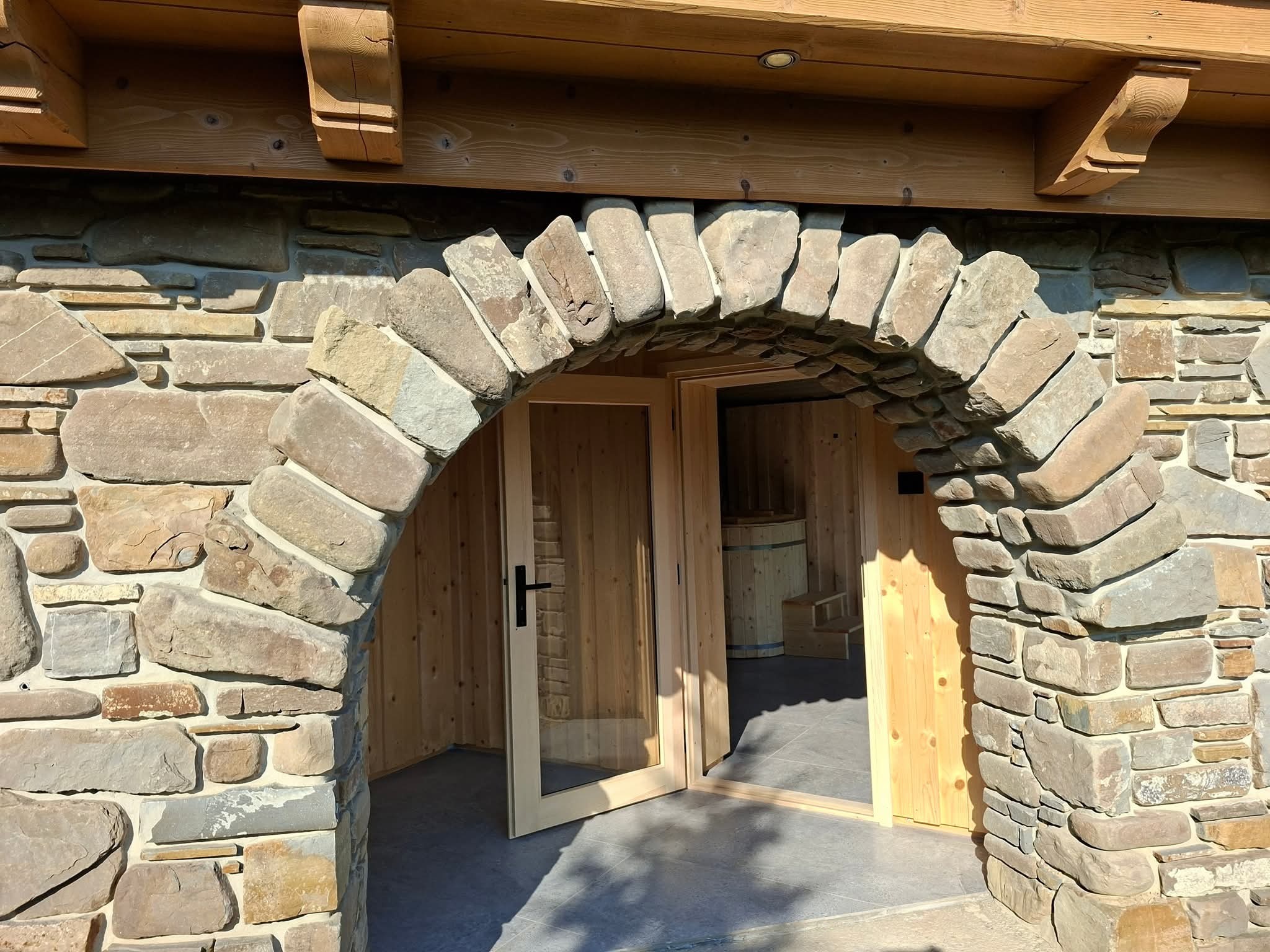 Kamienny łuk wejściowy do drewnianej sauny z beczką w środku, widok przez otwarte, przeszklone drzwi. Naturalne światło podkreśla teksturę kamienia i drewna.