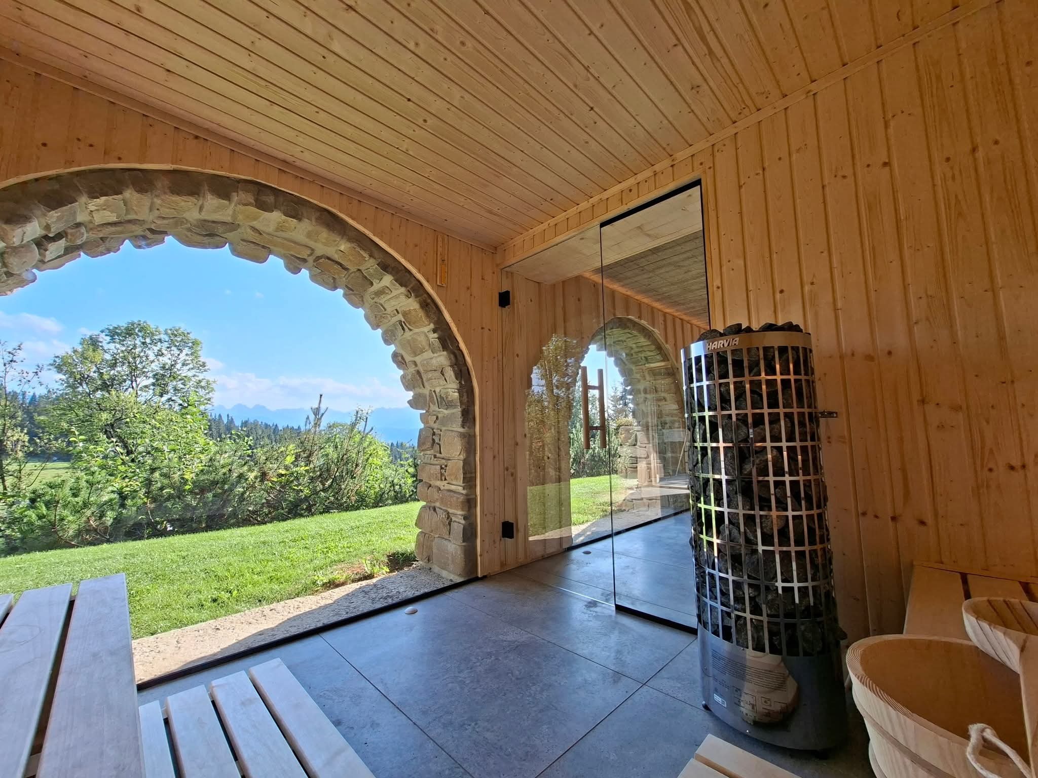 Wnętrze sauny z drewnianym wykończeniem, piecem Harvia i widokiem na góry przez kamienny łuk. Relaks w luksusowym otoczeniu.