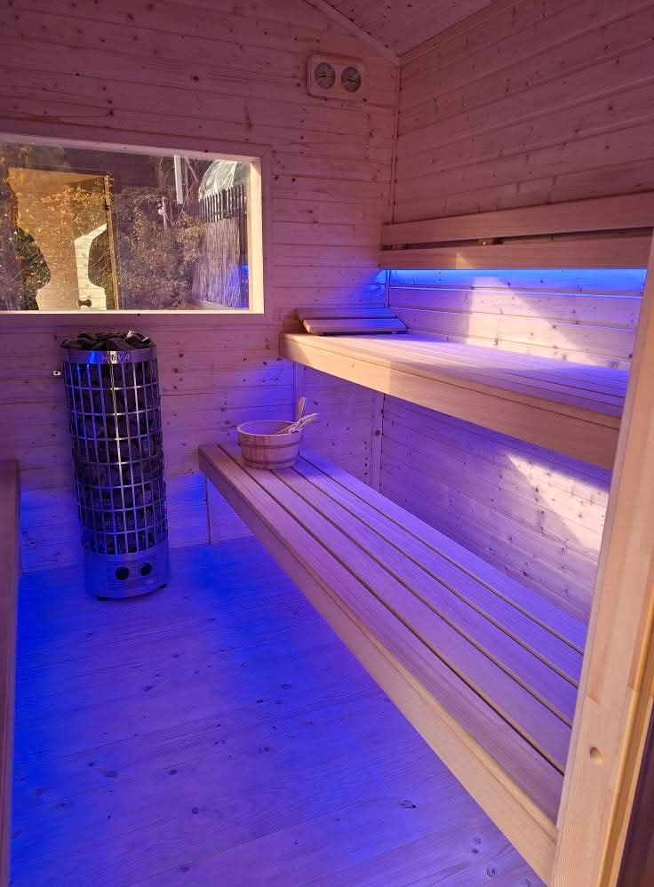 Jasne drewno i niebieskie podświetlenie w saunie: piec Harvia, okno z widokiem na zewnątrz, drewniane ławki i akcesoria do sauny. Przytulna atmosfera.