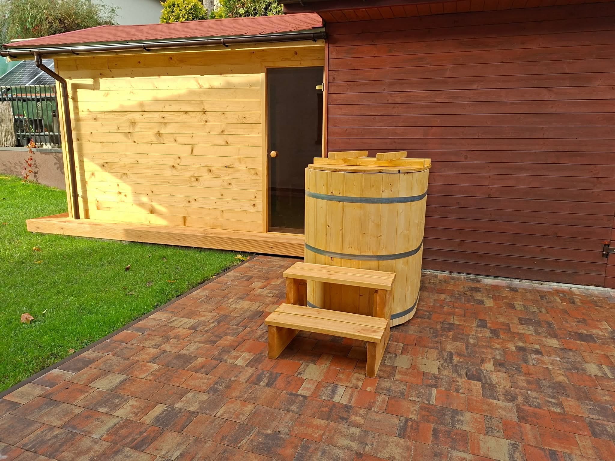 Drewniana sauna z beczką do schładzania i schodkami na zewnątrz, na tle brązowej ściany i kostki brukowej. Widok na drzwi sauny i fragment dachu.